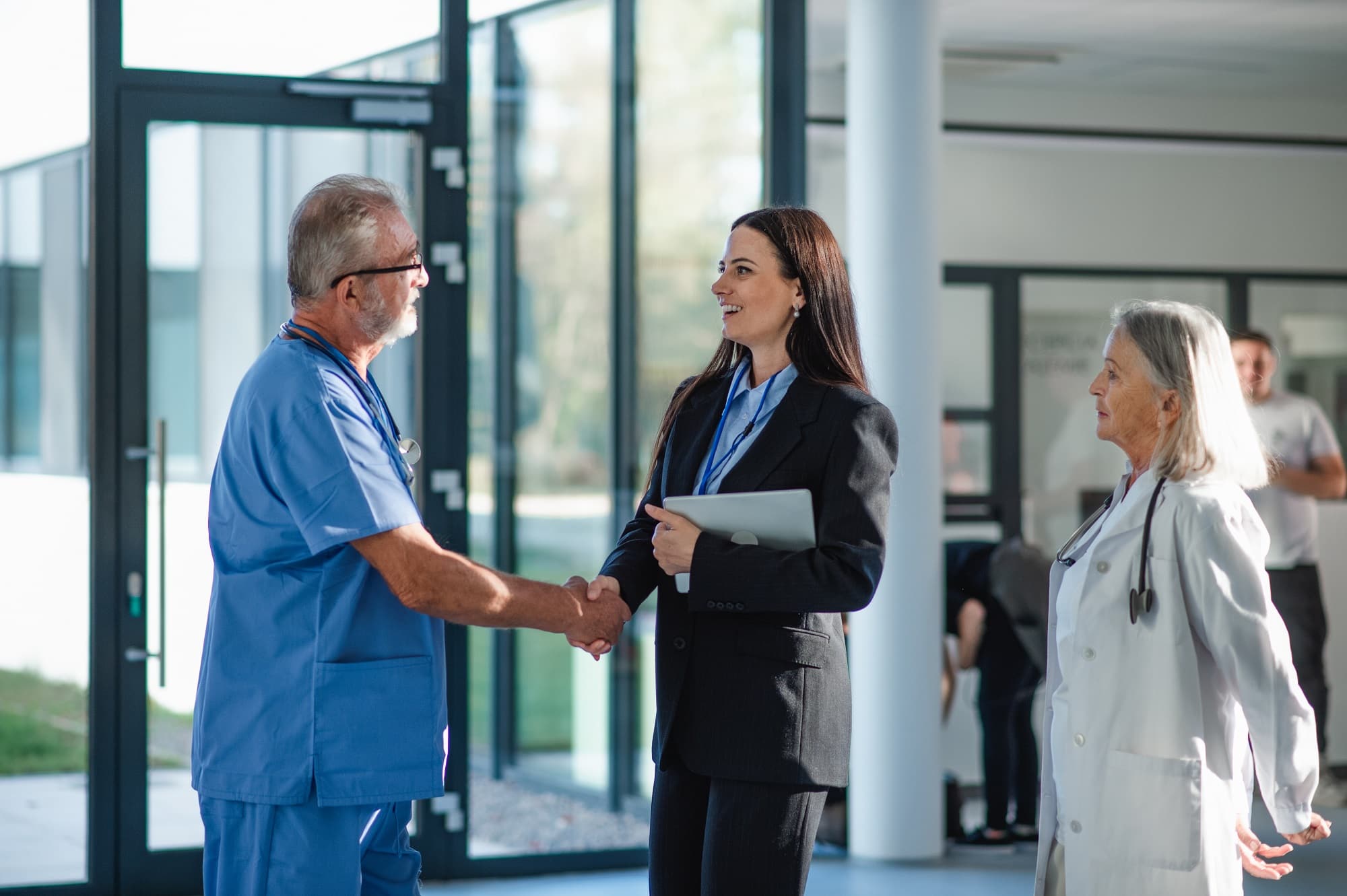 Photo of a woman in business attire shaking hands with a man in blue scrubs and a woman in a white lab coat.